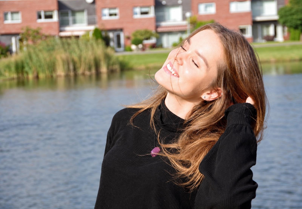 A woman in a black top wears a purple pendant necklace, smiling outdoors by water with houses in the background.