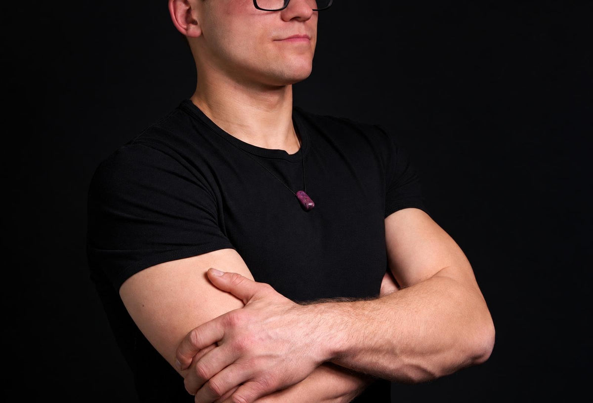 A man in a black t-shirt wears a dark purple pendant on a thin cord against a dark background.