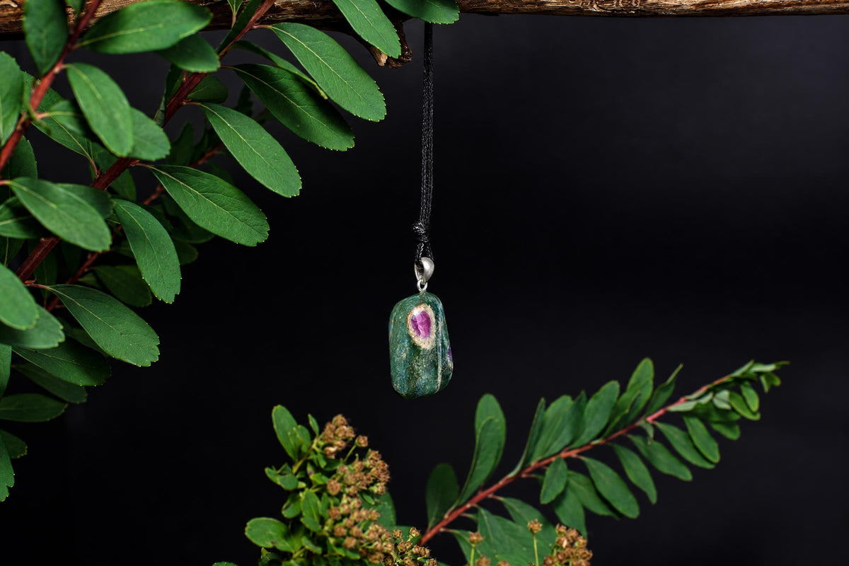 A green and purple stone pendant hangs from a black cord against a dark background, framed by green leaves.