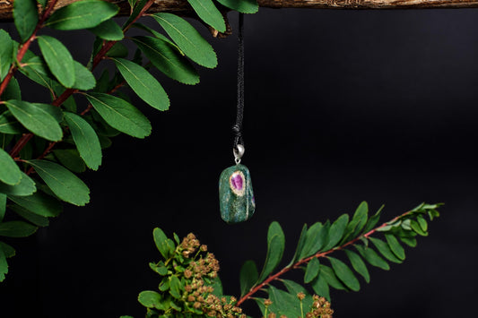 A green and purple stone pendant hangs from a black cord against a dark background, framed by green leaves.