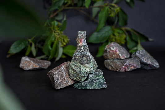 Several rough, green and brown speckled stones with pink inclusions are arranged on a dark surface near green leaves.