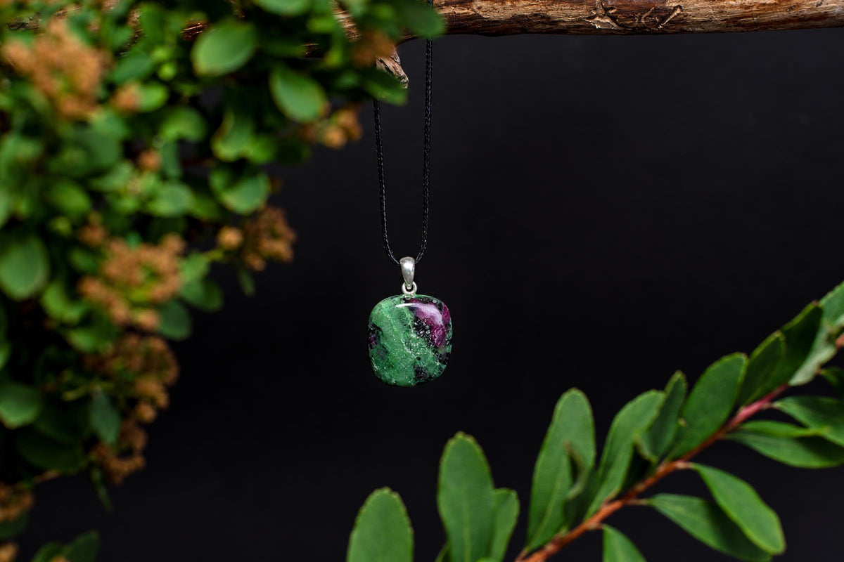 A polished green and purple stone pendant on a black cord, hanging from a branch against a dark background.