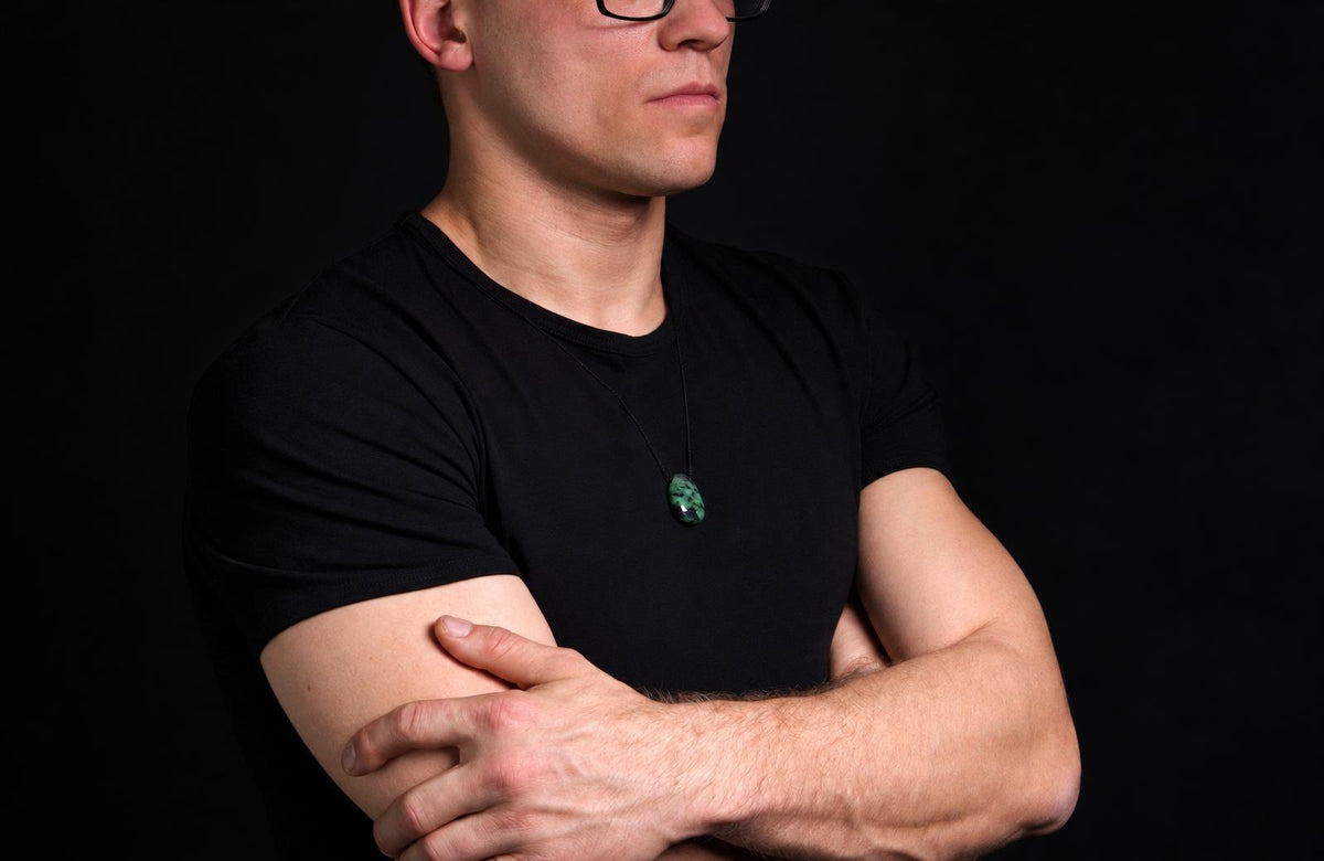 A man in a black t-shirt wears a dark green pendant with lighter speckles on a thin cord against a dark background.