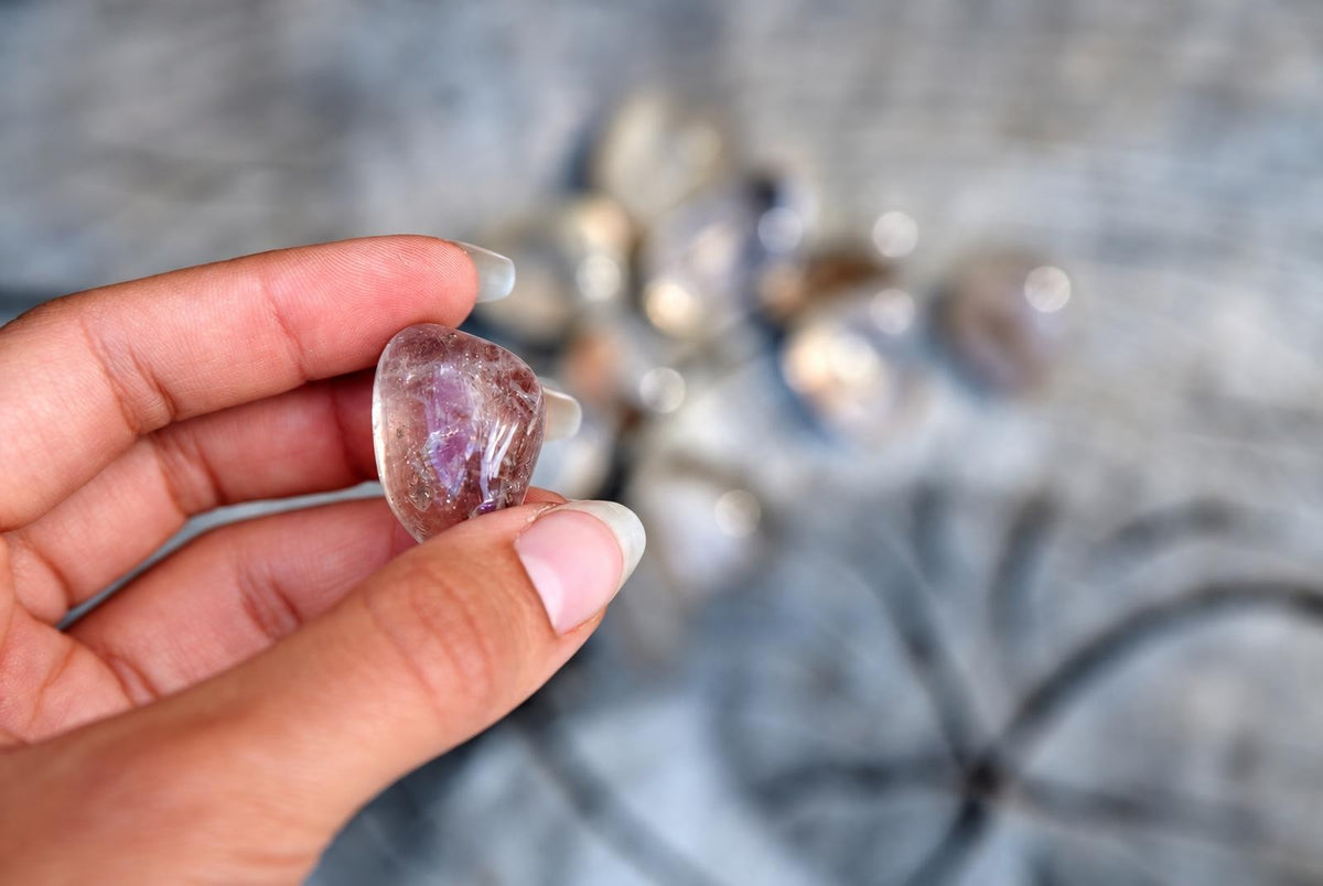 A hand holds a polished, translucent Rutile quartz stone with purple and brown inclusions against a blurred grey surface.
