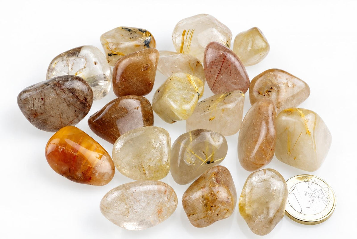 A collection of polished rutile quartz stones in various shades of brown, yellow and clear with golden needle-like inclusions, arranged on a white surface next to a coin.