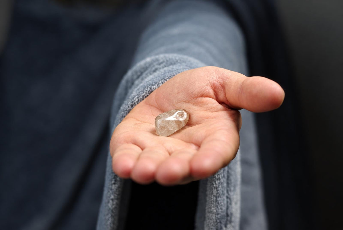A hand holds a small, translucent Rutile quartz stone with visible internal inclusions against a dark surface.