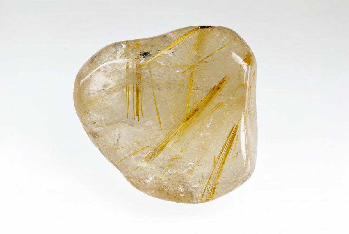 A polished, translucent Rutile quartz specimen with golden needle-like inclusions inside a pale yellow stone on a white surface.