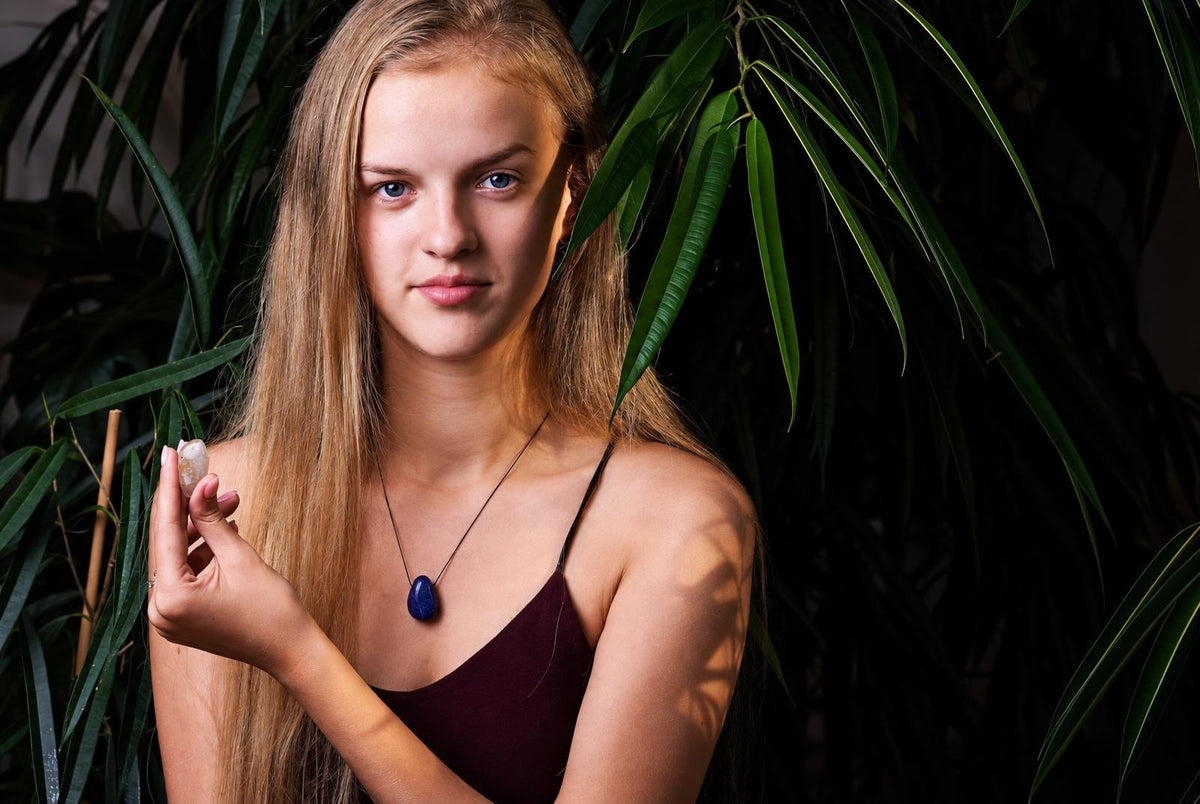 A woman with long blonde hair holds a small, pale stone in her hand. She wears a dark top and a necklace with a blue pendant.