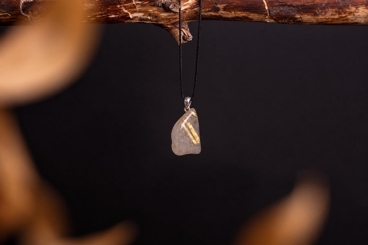 A clear, irregularly shaped pendant with golden rutile inclusions hangs from a black cord on a dark surface.