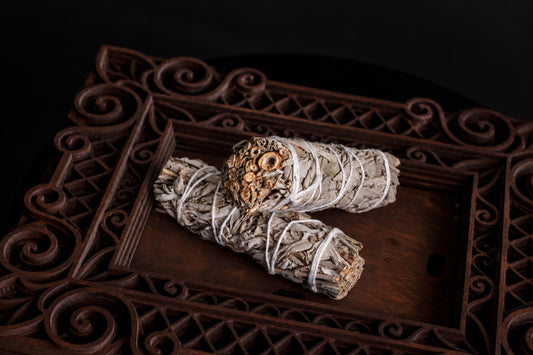 Two sage leaf incense sticks tied with white string rest on a dark wooden tray with ornate carved edges.