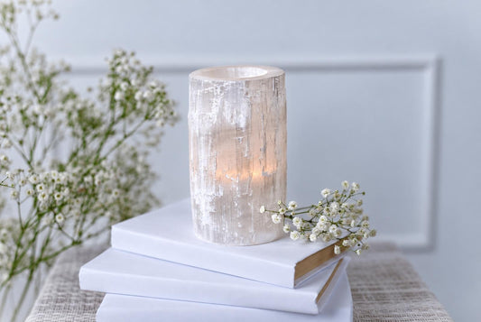 A cylindrical selenite candle holder with a textured, translucent surface sits on stacked white books beside small white flowers.