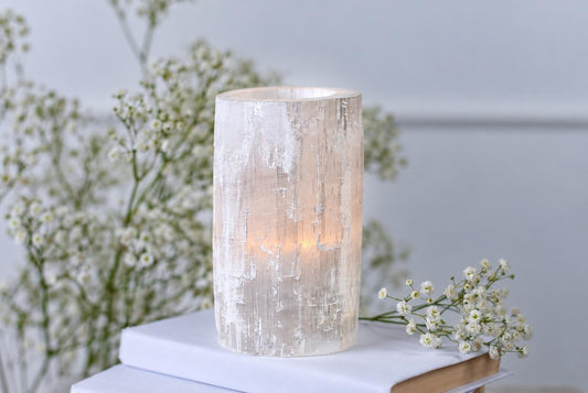 A cylindrical, translucent white candle holder with a textured surface sits on books beside small white flowers.