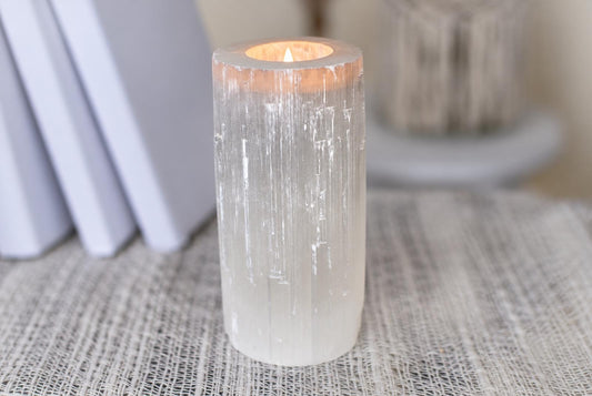 A cylindrical selenite candle holder with a lit flame inside, resting on a pale woven surface.