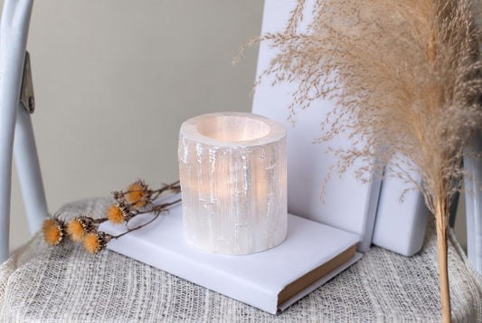 A cylindrical, translucent white selenite candle holder with a warm glow sits on a white book atop a textured surface.