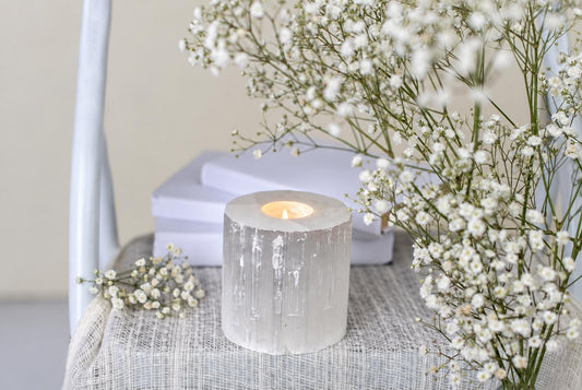A lit candle sits inside a translucent, cylindrical selenite holder with vertical striations on a pale woven surface.