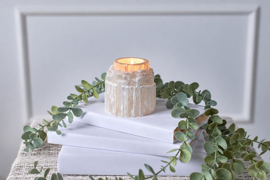 A cylindrical white stone candle holder with a glowing candle sits on stacked white books among trailing green leaves.