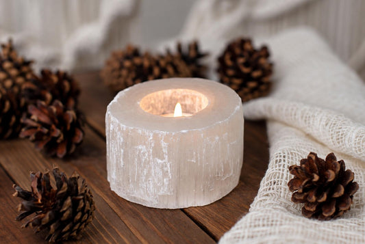 A lit candle inside a translucent, cylindrical selenite holder with vertical striations, surrounded by pinecones on a dark wooden surface.