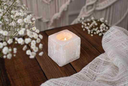 A square, pale candle holder with a lit tea light sits on a dark wooden surface beside white flowers and fabric.