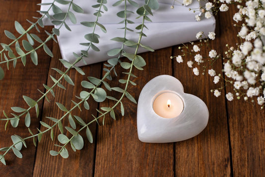 A white heart-shaped stone candle holder with a lit tealight rests on wooden boards among trailing green leaves and small white flowers.