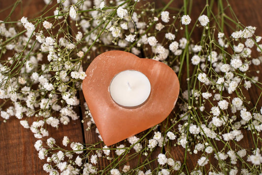 A heart-shaped, pinkish-orange candle holder with a white tea light sits on a dark wooden surface surrounded by small white flowers.