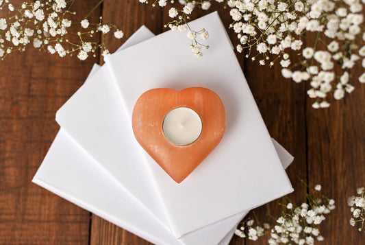 A heart-shaped, peach-coloured candle holder with a white tea light sits on white paper atop a dark wooden surface, surrounded by small white flowers.