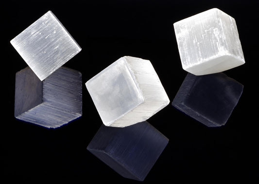 Three translucent white selenite cubes with visible striations rest on a dark reflective surface against a black background.