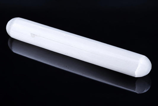 A white, cylindrical selenite massage stick with rounded ends rests on a dark, reflective surface.