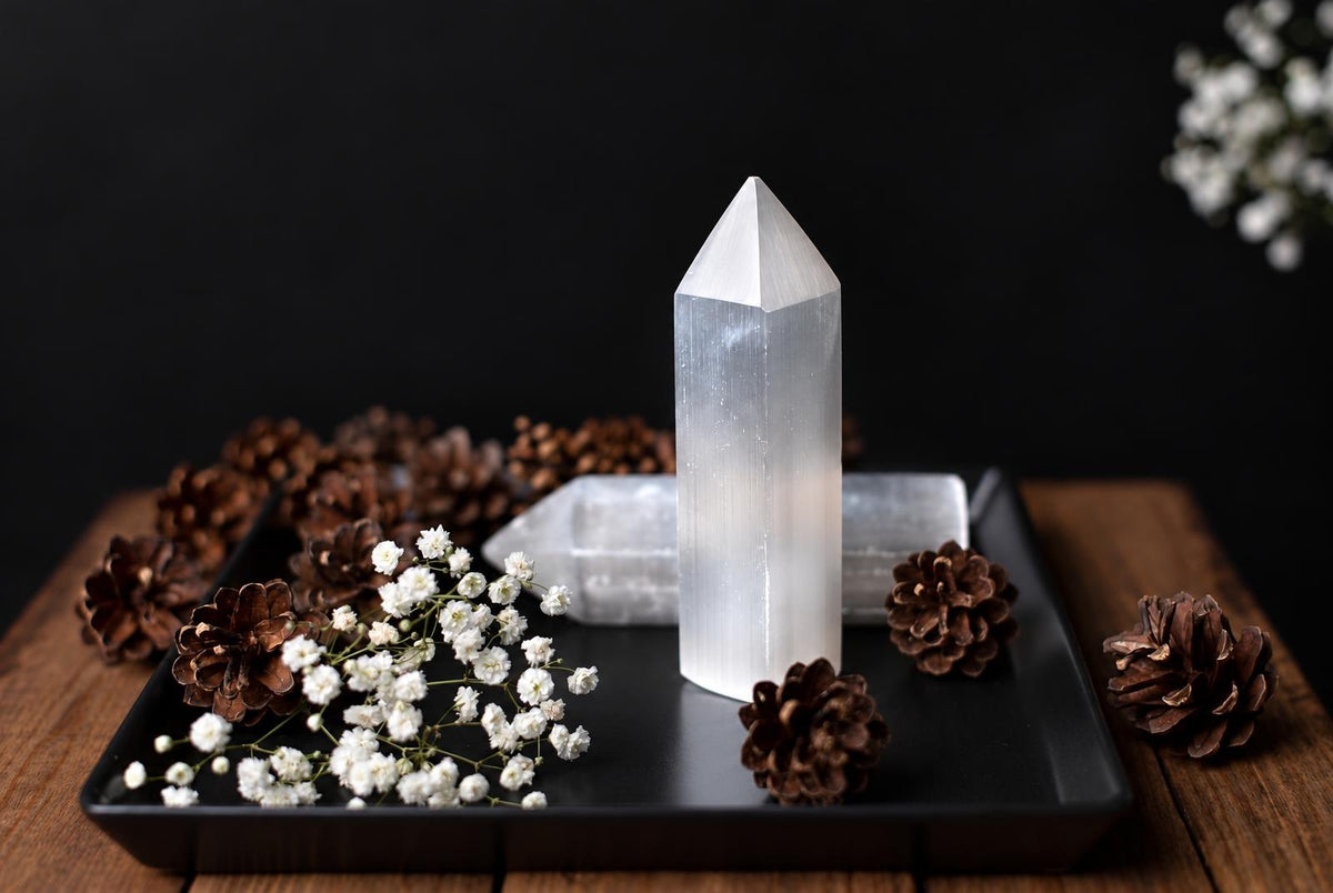 A white pointed selenite obelisk stands on a black tray beside brown cones and small white flowers.