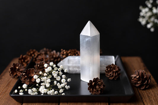 A white pointed selenite obelisk stands on a black tray beside brown cones and small white flowers.