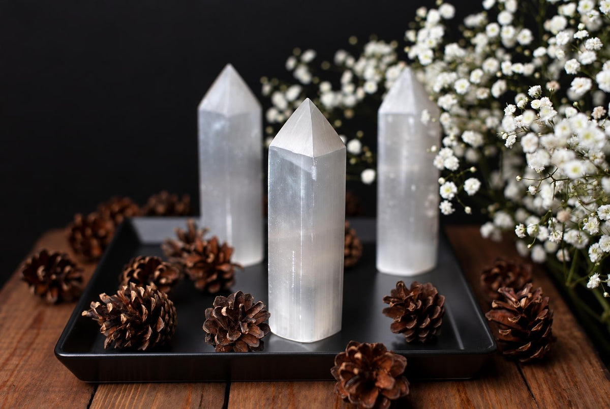 White pointed selenite towers stand on a black tray among brown cones and small white flowers.