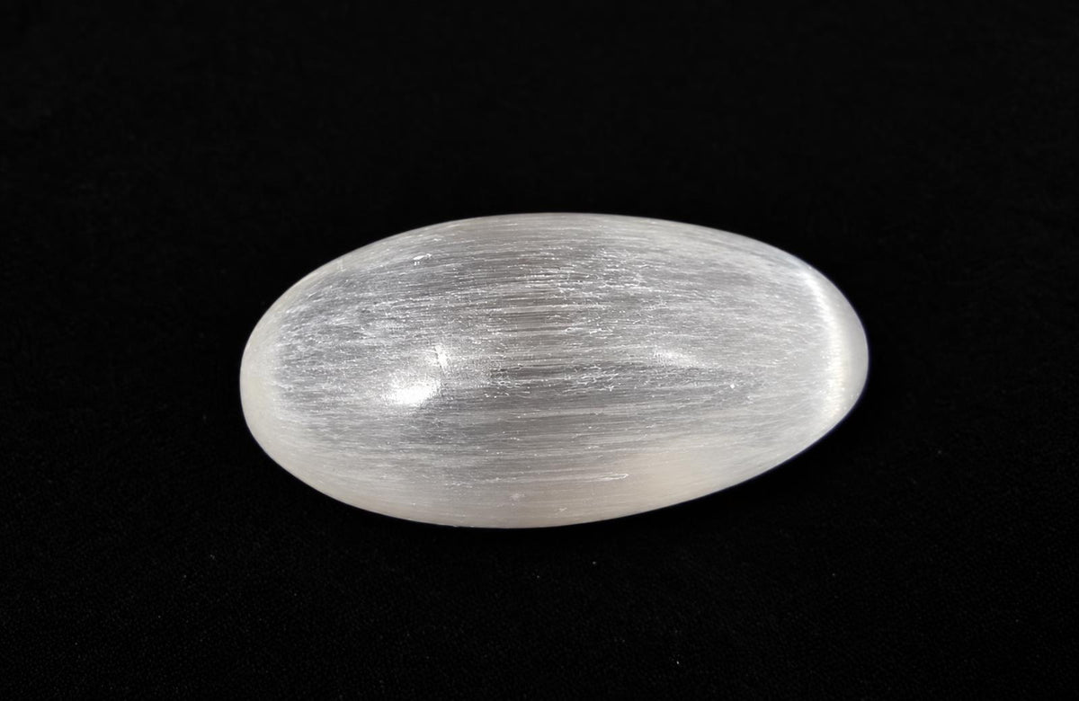 A polished, oval-shaped selenite palm stone with a white, fibrous texture on a dark surface.