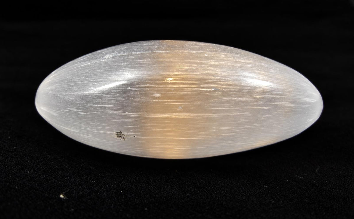 A polished, oval-shaped selenite palm crystal with a smooth white surface and golden-brown striations on a dark background.