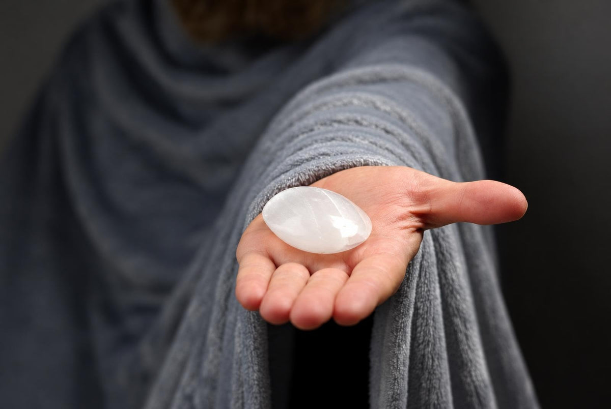 A person in a grey robe holds a smooth, white, oval-shaped polished stone in their open palm against a dark background.