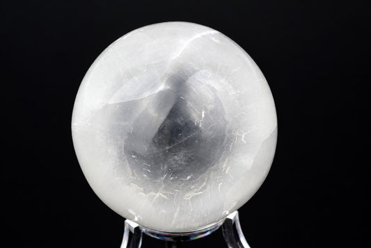 A polished, translucent white sphere with internal grey veining sits on a clear stand against a dark background.