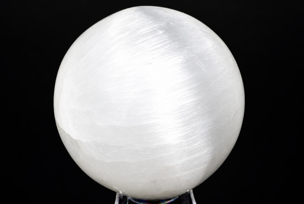 A white, translucent selenite sphere with a smooth surface sits on a clear stand against a dark background.