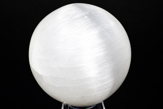 A white, translucent selenite sphere with a smooth surface sits on a clear stand against a dark background.