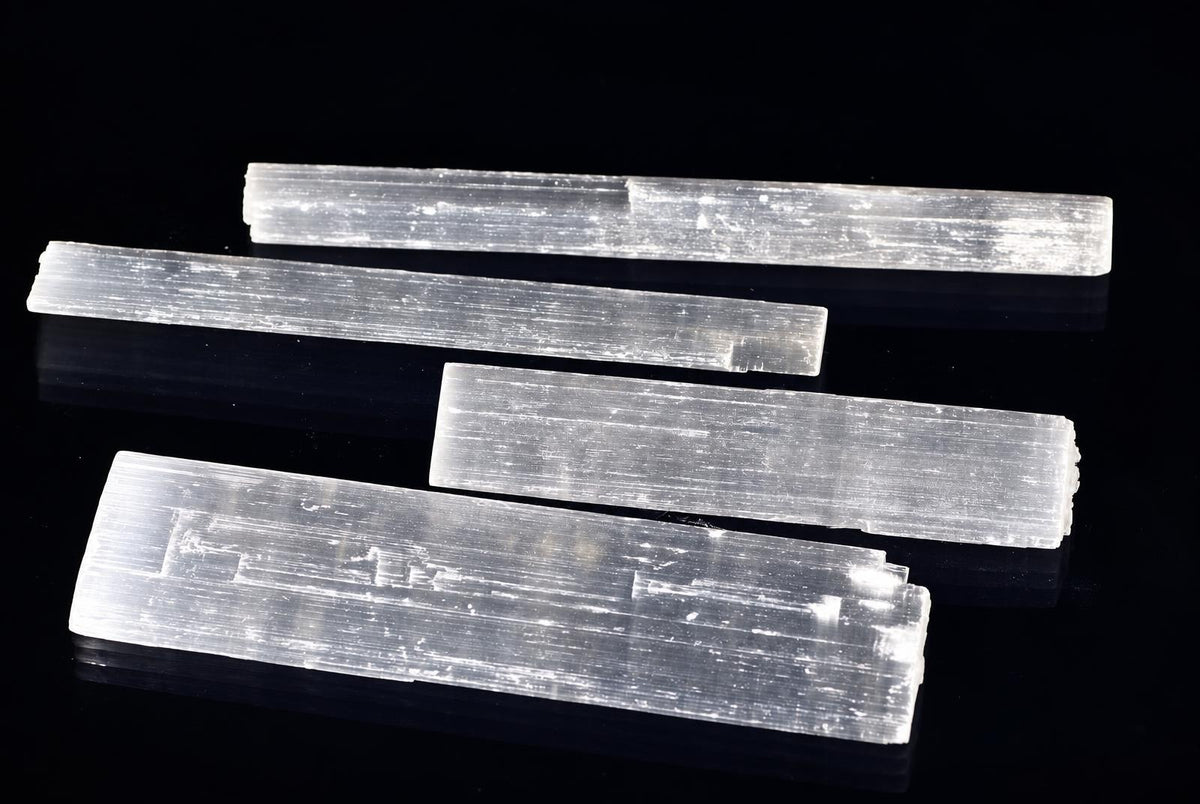Four translucent, rectangular selenite sticks with a striated texture lie on a dark surface.