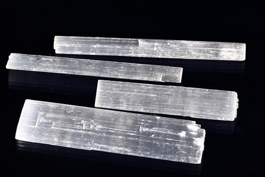 Four translucent, rectangular selenite sticks with a striated texture lie on a dark surface.