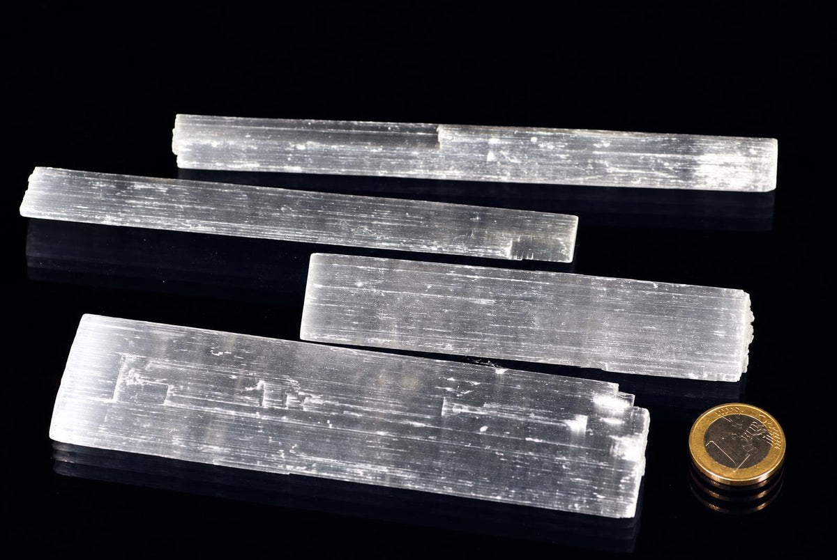 Four pale translucent selenite sticks show long parallel striations and flat rectangular shapes, arranged beside a coin on a reflective black background.