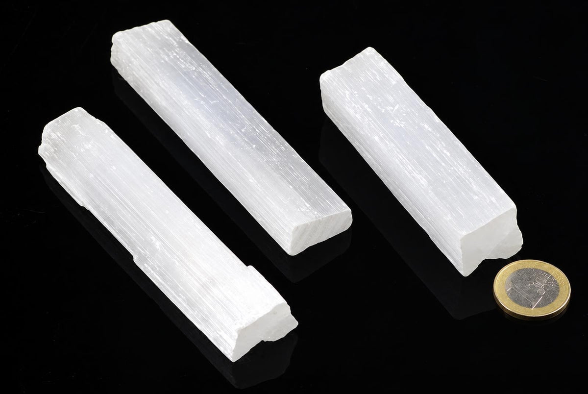 Three white, rectangular selenite sticks with a fibrous texture lie on a dark surface next to a coin.