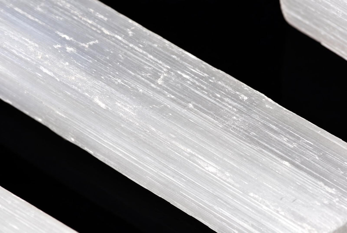 A close-up of a translucent white selenite stick with visible striations, resting on a dark surface.