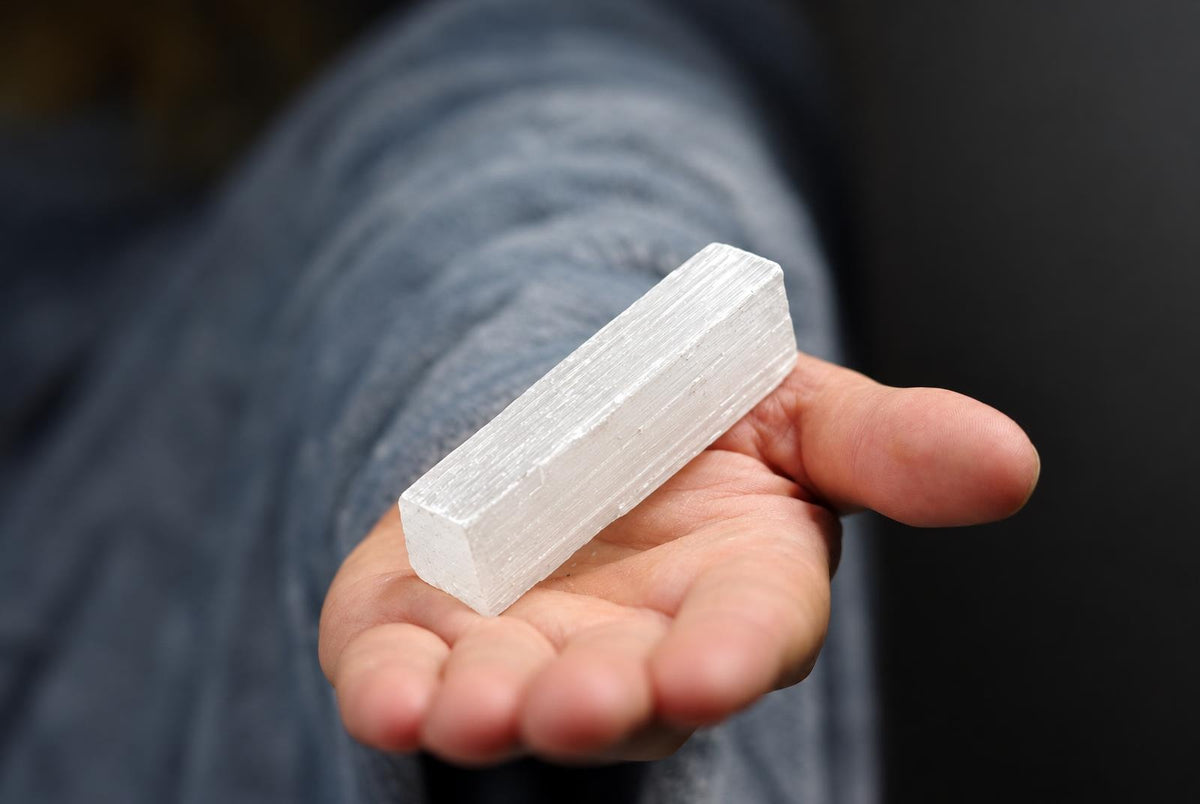 A hand holds a white, rectangular selenite stick with visible striations against a dark background.