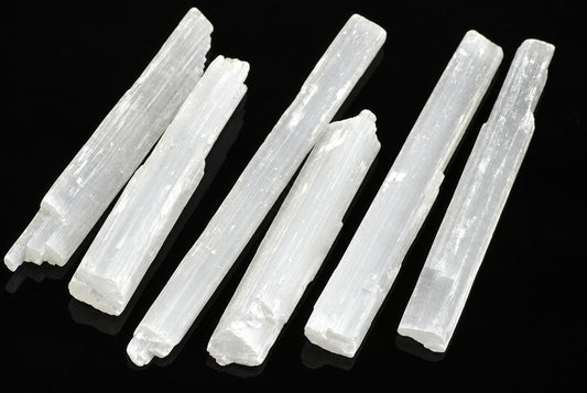 Six white, translucent selenite sticks with vertical striations lie on a dark surface.