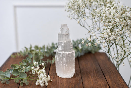 A tall, translucent selenite crystal tower with stepped facets stands on a dark wooden surface beside white flowers and greenery.