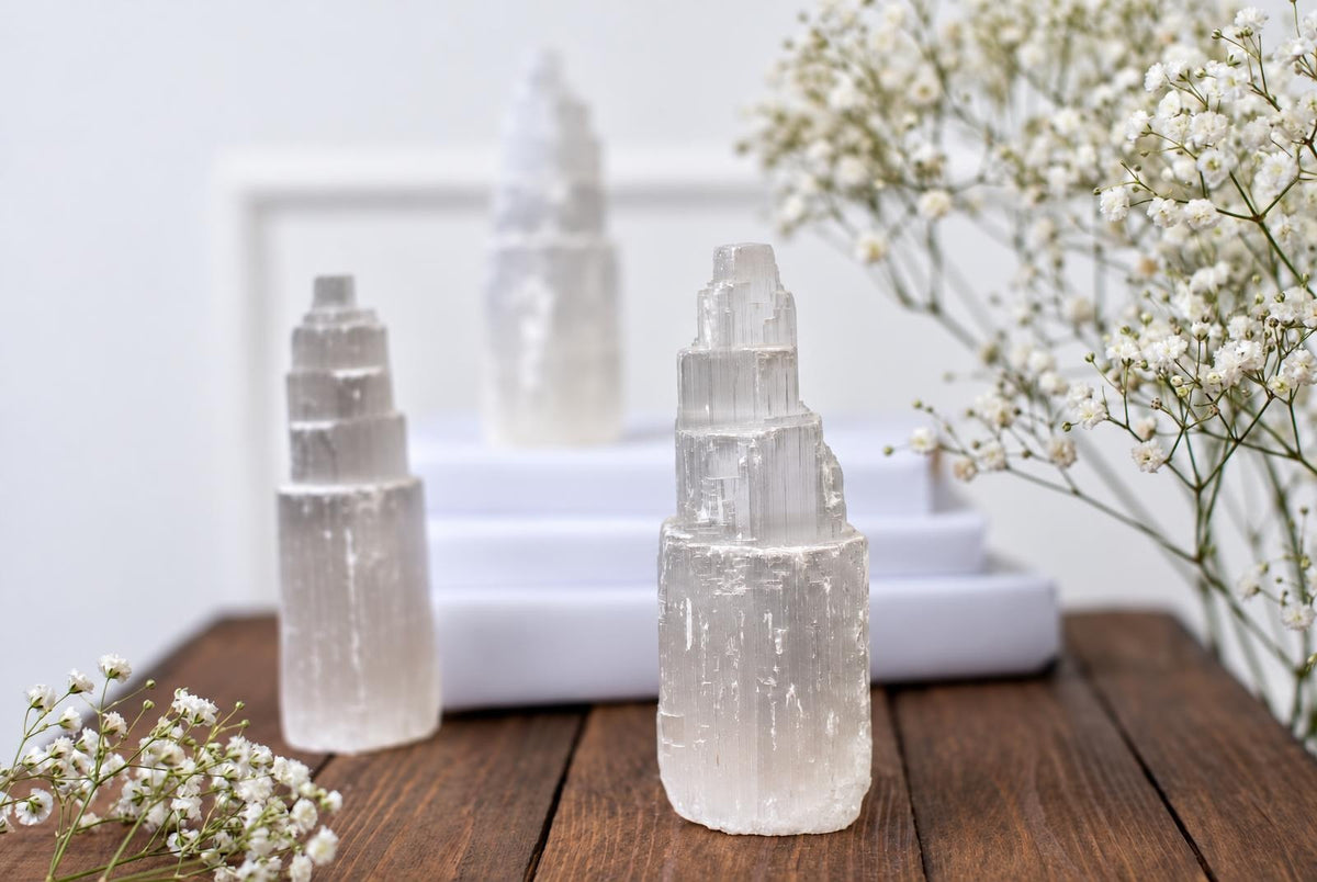 Three translucent, tiered selenite towers stand on a dark wooden surface beside white flowers.