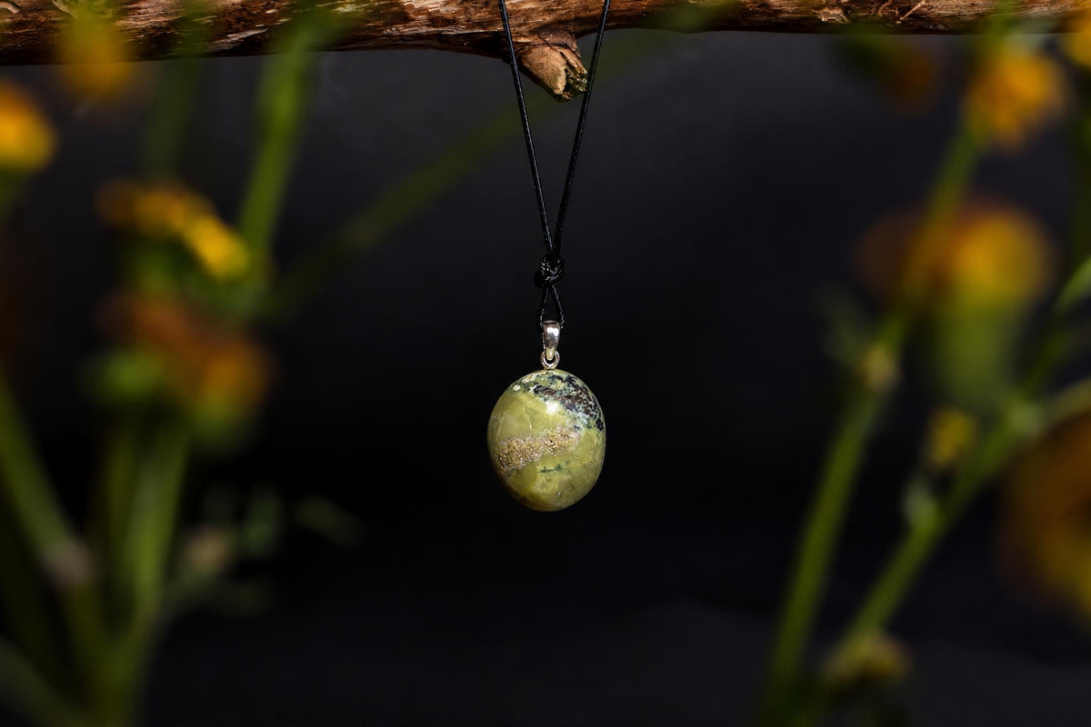A polished oval pendant with green and brown patterns hangs from a black cord on a dark surface.