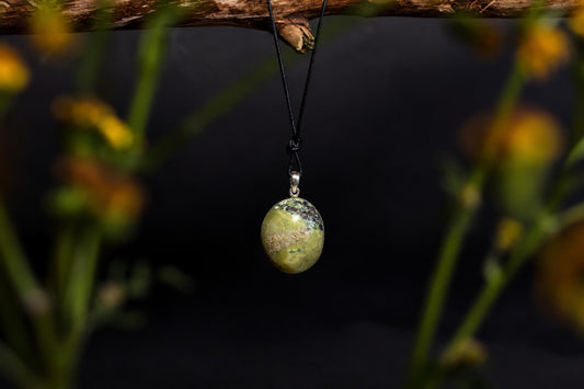 A polished oval pendant with green and brown patterns hangs from a black cord on a dark surface.