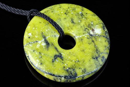 A yellow-green donut-shaped pendant with black and white speckles, a central hole, and a dark cord against a dark surface.