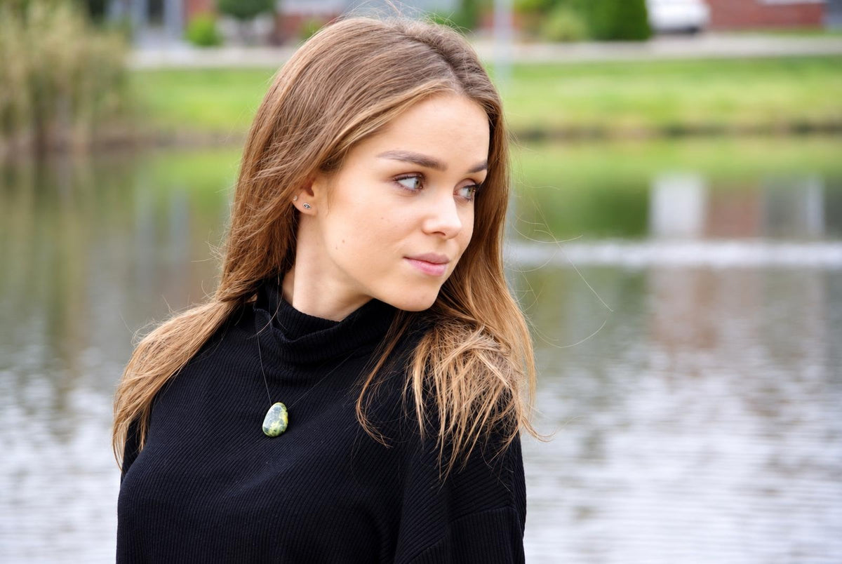 A woman wears a black turtleneck with a green and white patterned oval pendant on a thin chain, set against a blurred water background.