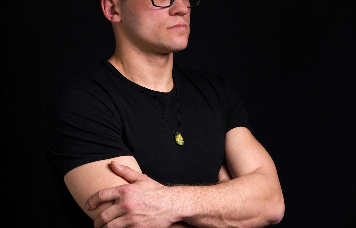 A man in a black t-shirt wears a pendant with an oval, yellow-green stone on a dark cord against a black background.